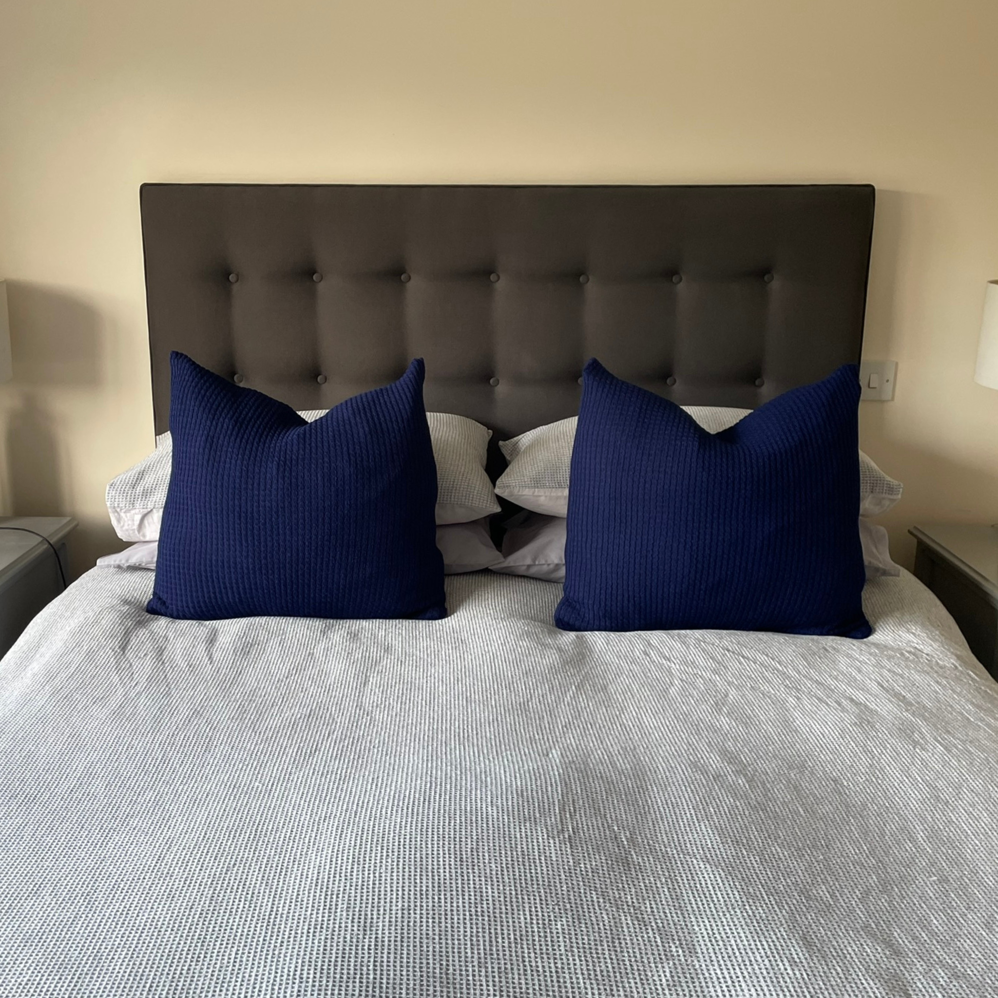 Two large cushions with navy blue alpaca cushion covers on a bed with grey bedding.