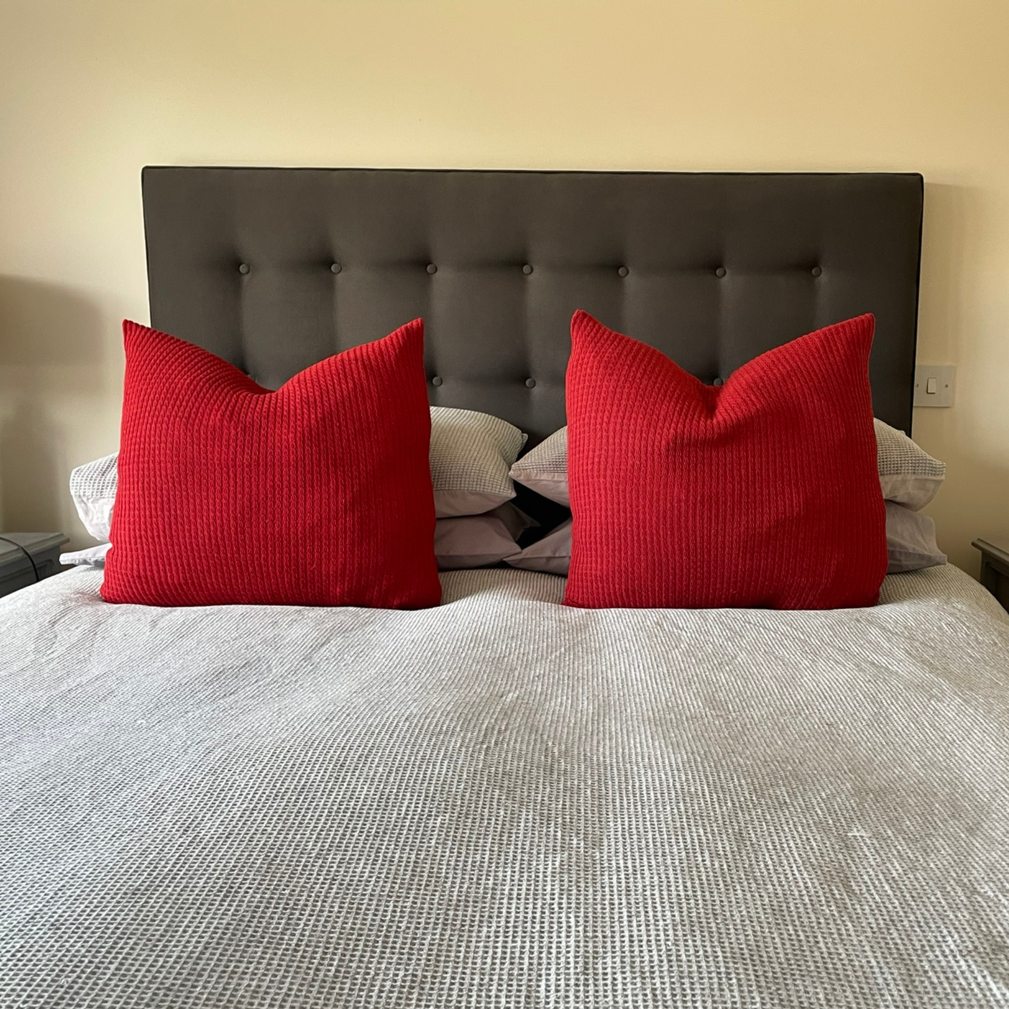 Two large cushions with red alpaca cushion covers on a bed with grey bedding.