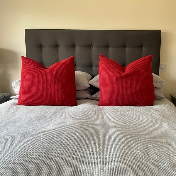 Two large cushions with red alpaca cushion covers on a bed with grey bedding.