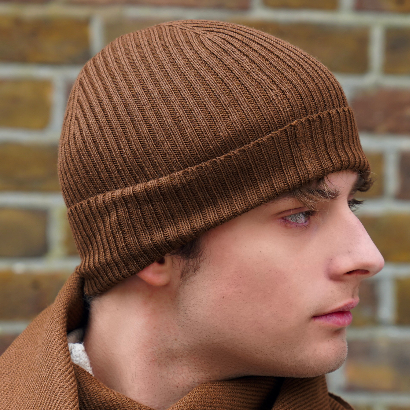 Young man wearing brown alpaca beanie hat in front of brick wall in outdoor setting.