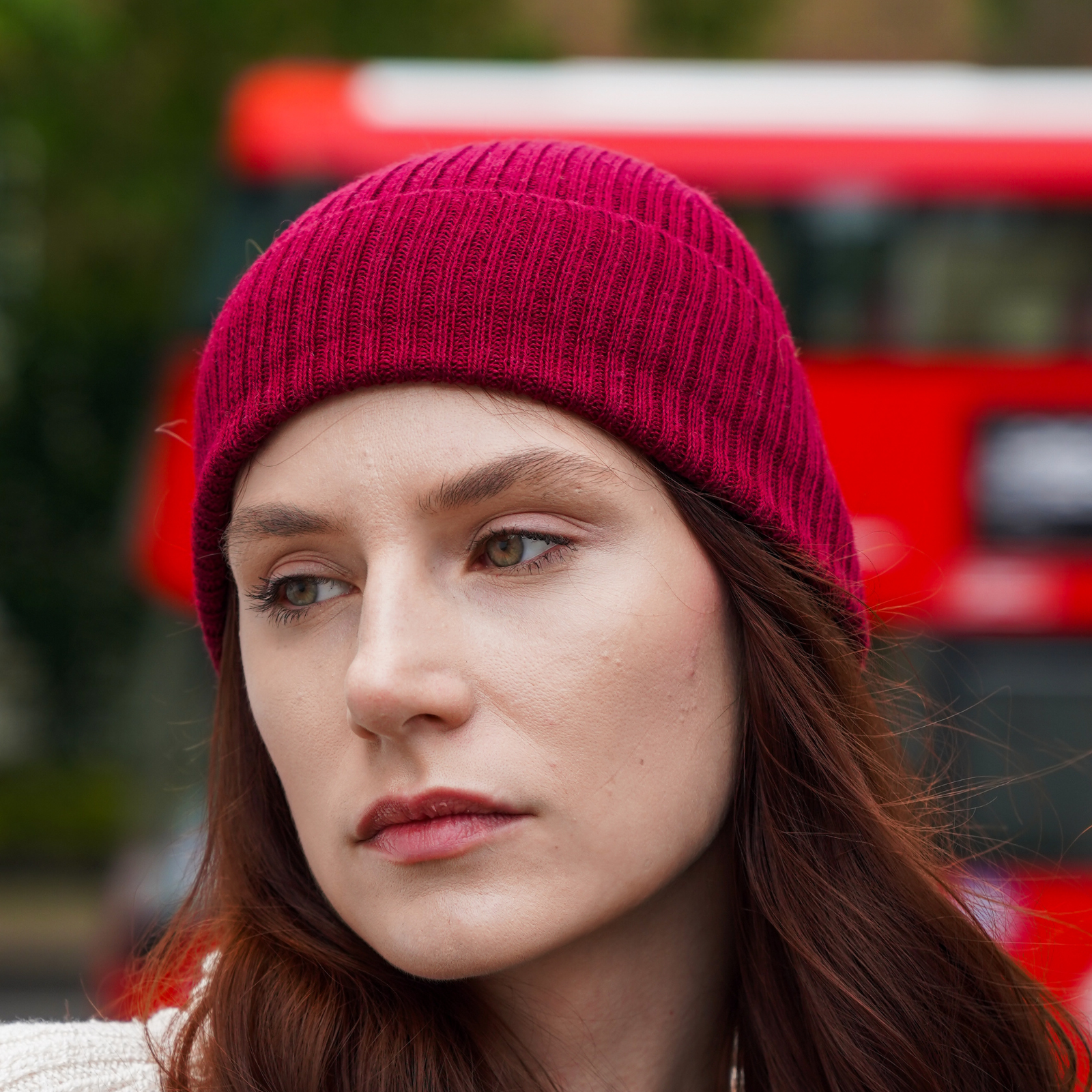 Young woman wearing Burgundy alpaca beanie hat in an outdoor setting in a city with a bus.