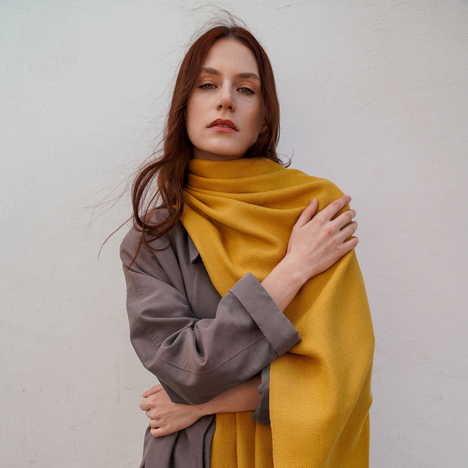 Young woman wearing camel colour long extra large alpaca wool scarf shawl in front of a wall.
