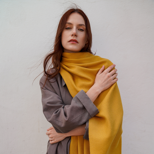 Young woman wearing camel colour long extra large alpaca wool scarf shawl in front of a wall.