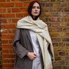 Young woman wearing cream long extra large alpaca wool scarf shawl in front of a brick wall