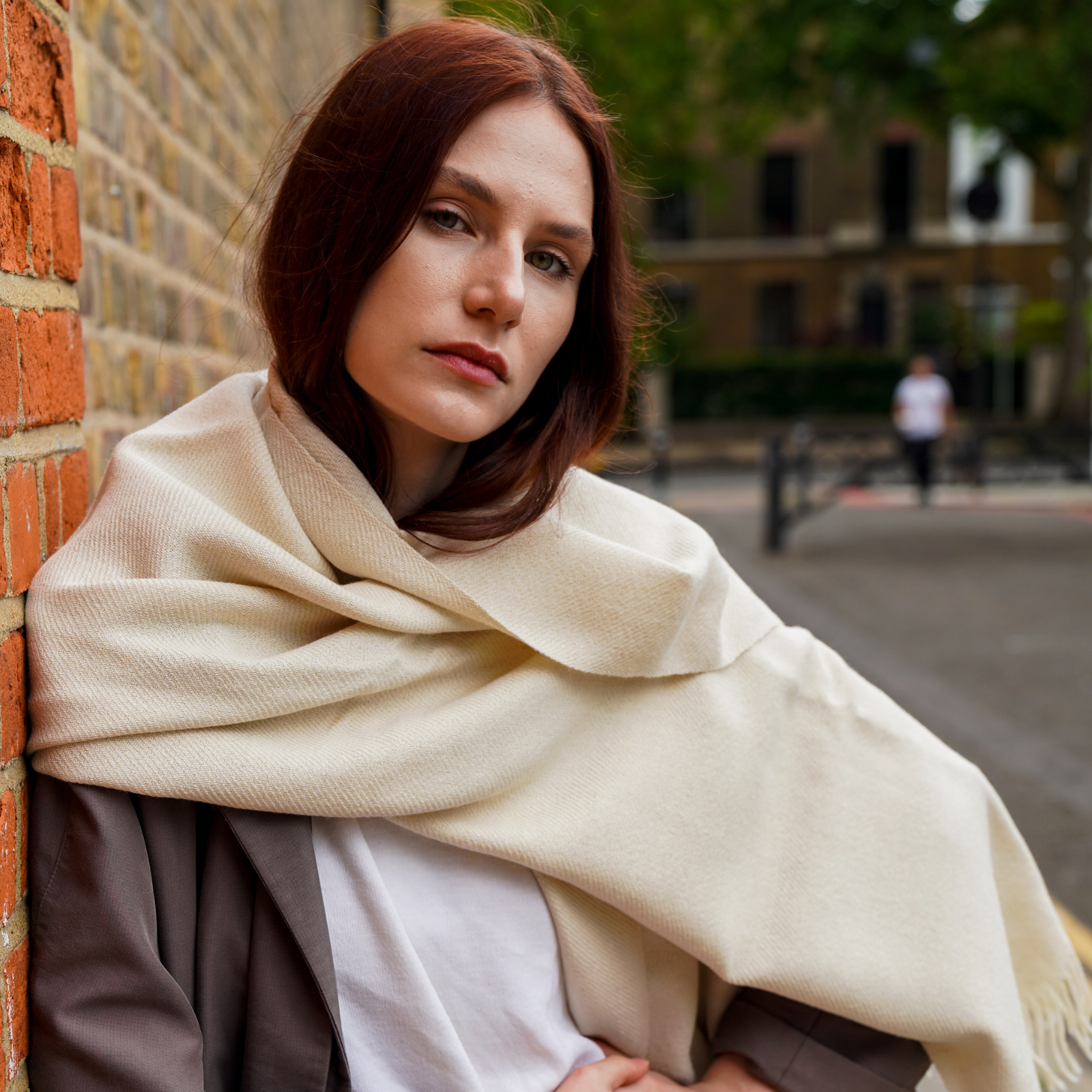 Young woman wearing cream long extra large alpaca wool scarf shawl in front of a brick wall in the city outdoors.